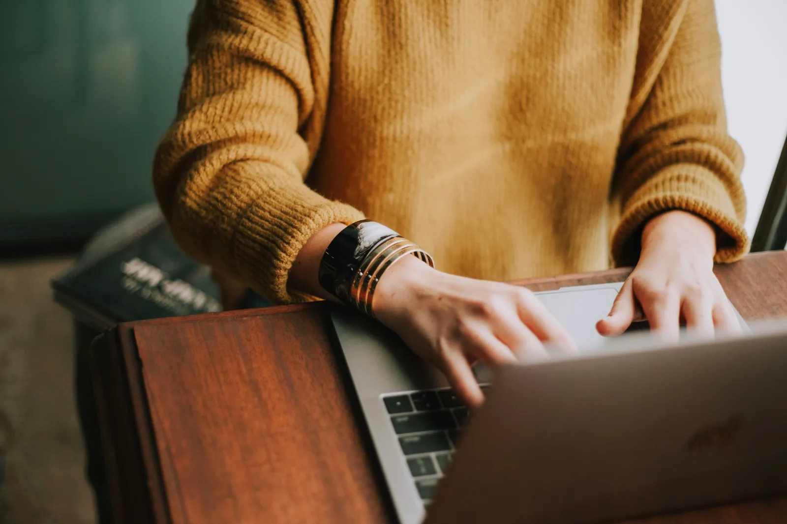 Someone in a mustard sweater typing on a laptop at a wooden desk